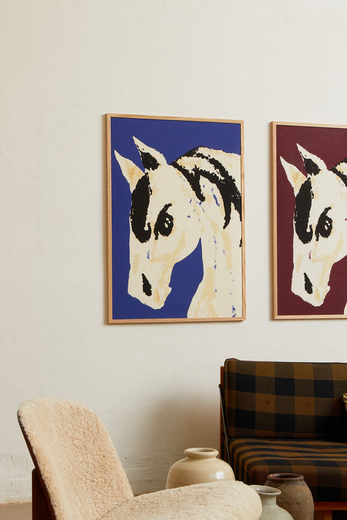 Art print with a stylized illustration of a horse head in light beige and black tones against a cobalt blue background, with an intentionally blurred and painterly feel. Styled in a minimalist interior.