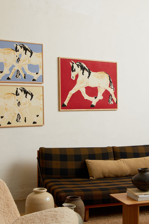 Art print with a stylized illustration of a horse in motion in light beige and black tones against a red background, with an intentionally blurred and painterly feel. Styled in a minimalist interior.