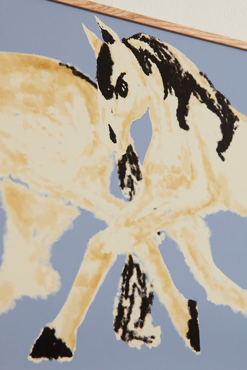 Art print with a stylized illustration of two mirrored horses in motion in light beige and black tones against a gray-blue background, with an intentionally blurred and painterly feel. Styled in a minimalist interior.