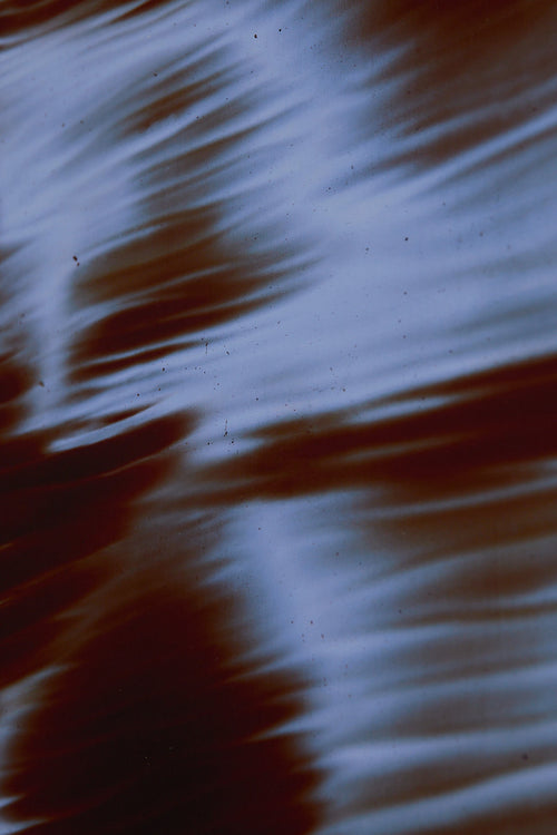 Art print featuring soft, blurred contours and flowing light blue shapes emerging against a dark red-brown surface, creating an impression of motion and impermanence. Styled in a minimalist interior.