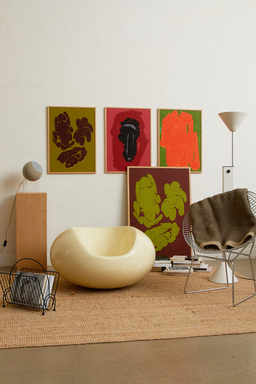 Art print featuring soft, blurred, and flowing bright orange shapes emerging against a dark green surface, creating an impression of motion and impermanence. Styled in a minimalist interior.