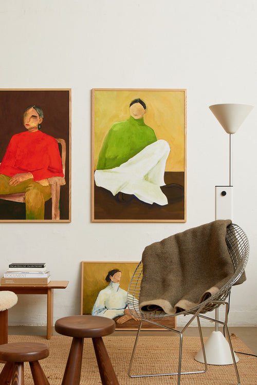 Art print featuring a painting of a person sitting on the ground, with a green sweater, light checkered bottoms, and dark loafers against a neutral background in dark brown and ochre. Styled in a minimalist interior.