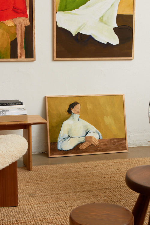 Art print featuring a painting of a person in a white chunky turtleneck against an olive-colored background, resting their hands on a wooden bench. Styled in a minimalist interior.