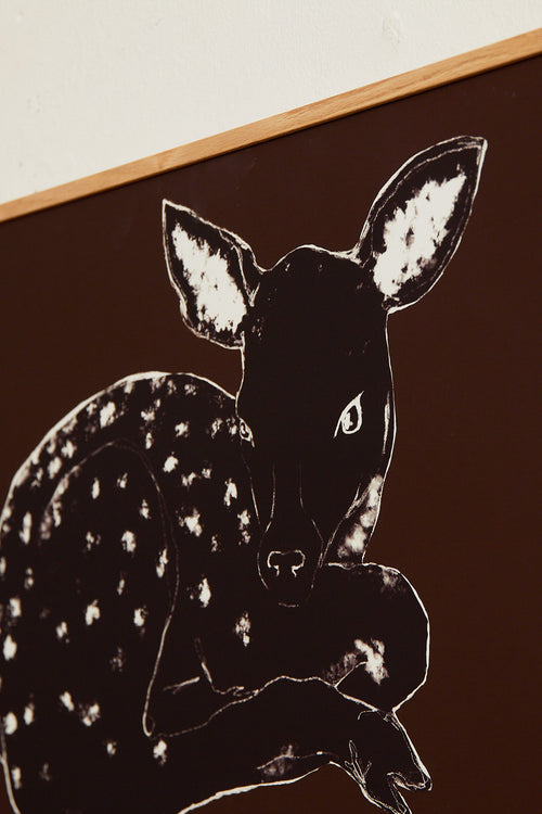 Art print with a stylized illustration of a lying deer in a dark tone against a dark brown background, with a minimalist and graphic expression. Styled in a minimalist interior.
