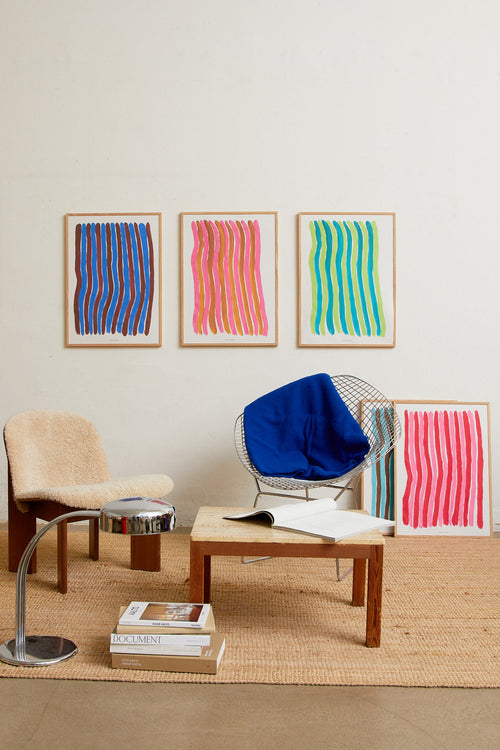Three abstract striped art prints in oak frames on a light wall, styled above a wooden coffee table with a chair and woven rug.