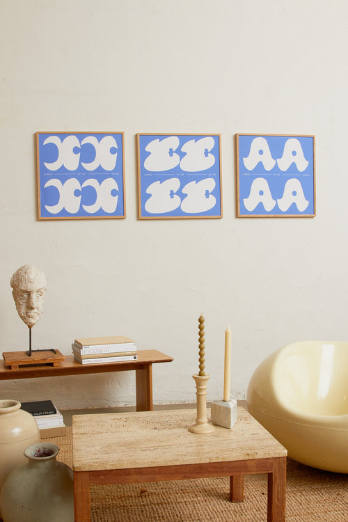 Square-format Art print with a graphic interpretation of the letter “E”. Created in white against a calm blue, lightly grain-textured background. Styled in a minimalist interior.