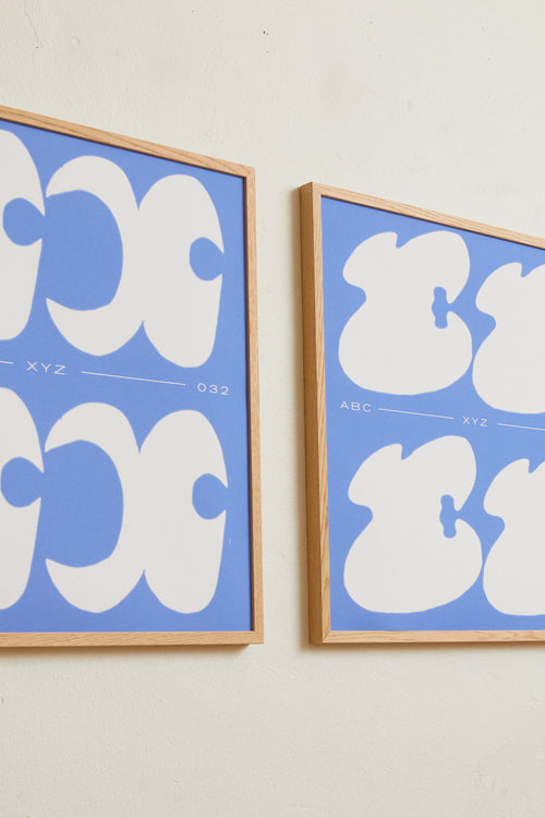 Square-format Art print with a graphic interpretation of the letter “X”. Created in white against a calm blue, lightly grain-textured background. Styled in a minimalist interior.