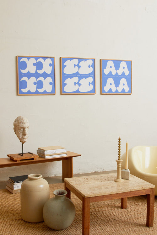 Square-format Art print with a graphic interpretation of the letter “X”. Created in white against a calm blue, lightly grain-textured background. Styled in a minimalist interior.