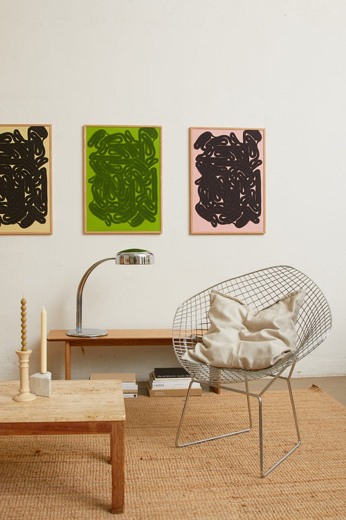 Art print featuring dense, black rounded forms with a smooth, glossy feeling emerging against a warm, light pink background. Styled in a minimalist interior.