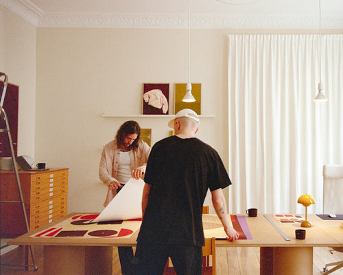 A man in a black t-shirt and white cap stands at a wooden table holding a large white sheet, while a woman in a beige cardigan works in the background of a bright, modern room with white curtains and wall art.