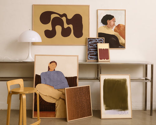 A styled interior scene showing multiple framed art prints and paintings leaning and hanging against a white wall beside a metal console table, lamp, stool, and small framed accessories.