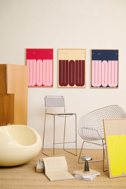 Three framed abstract art prints with bold wavy vertical stripes in pink, burgundy, and navy hang on a cream wall above a styled room scene with a stool, wire chair, books, and a bowl.